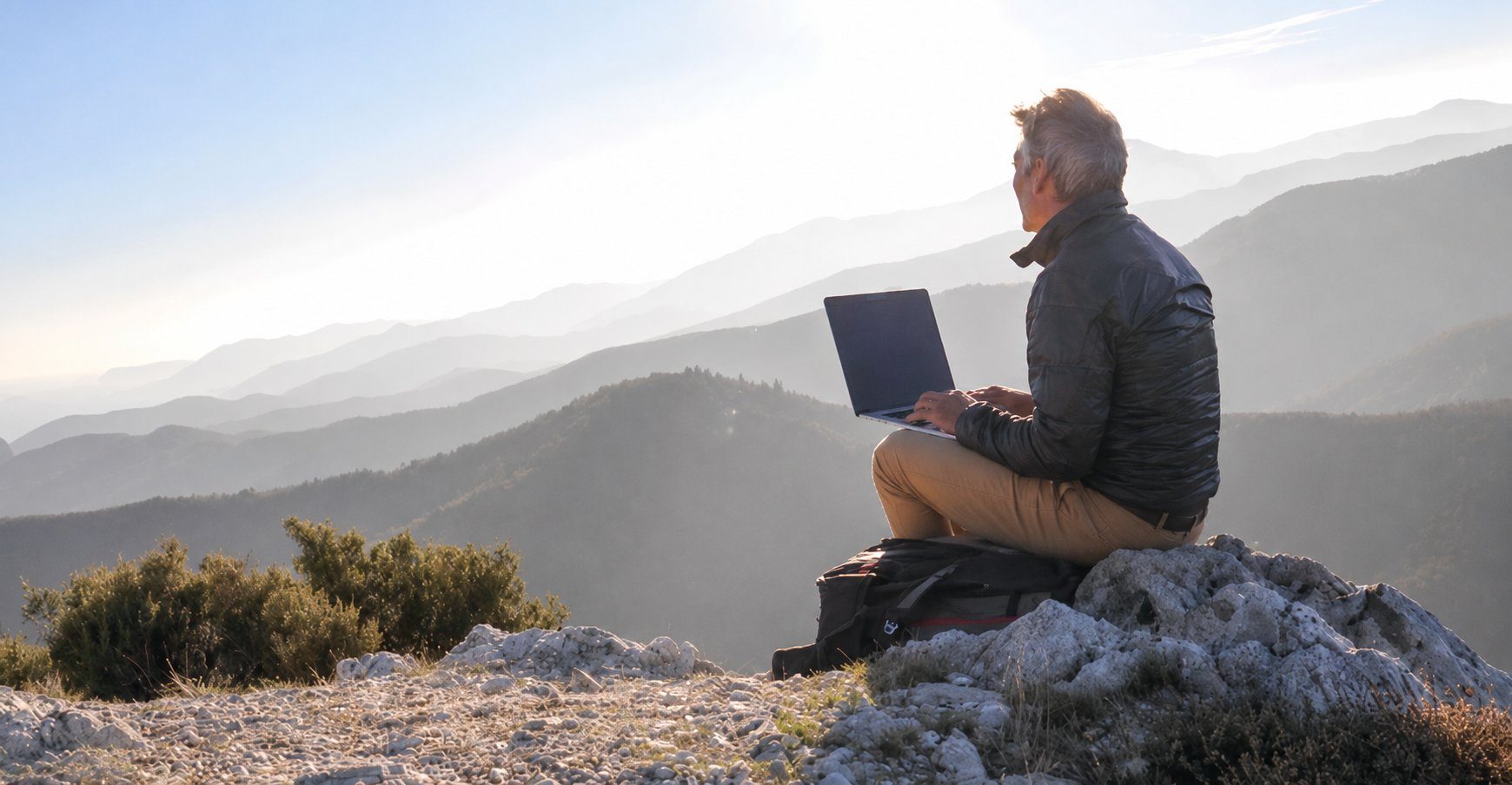 A person working on a laptop while sitting on a mountain summit at golden hour, looking out over layered mountain ranges
